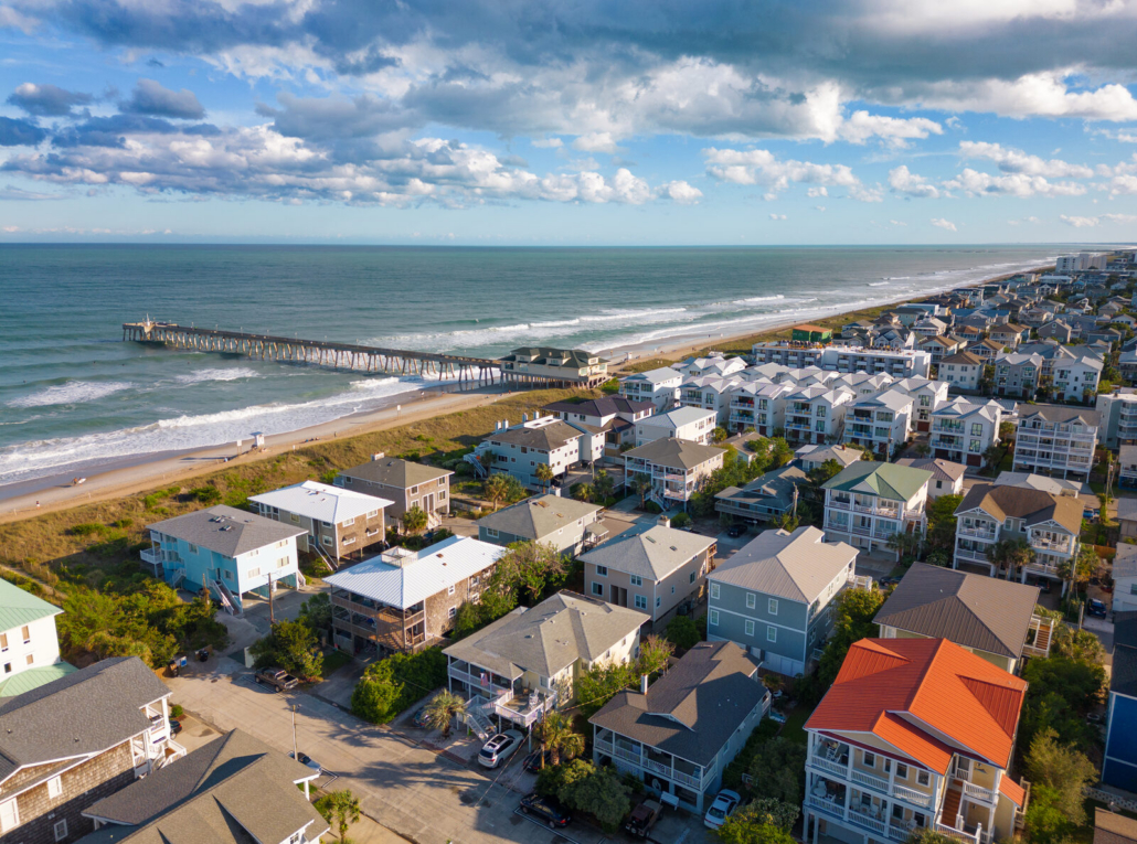 Rows of raised beach houses and their roofs along a shore street, with the beach and pier behind